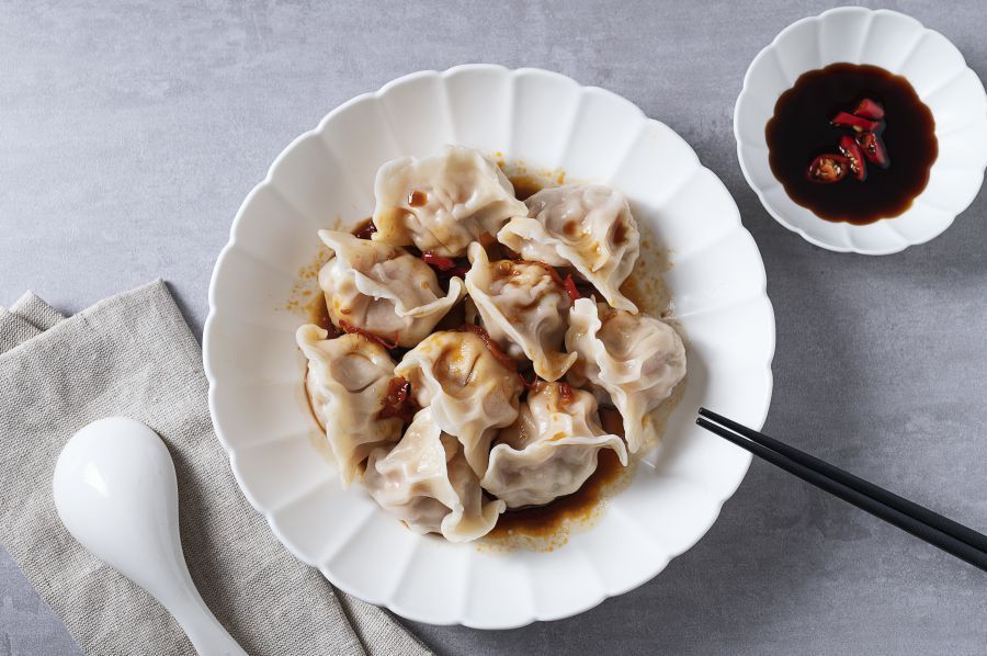 Shrimp dumpling held by black chopsticks, plate of dumplings, small dipping sauce, grey background.