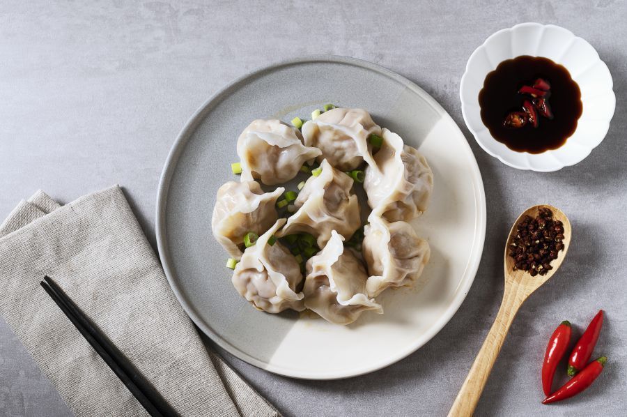 Dumplings on a plate, one being held by chopsticks with dipping sauce, ceramic bowl, and spoon in the background.