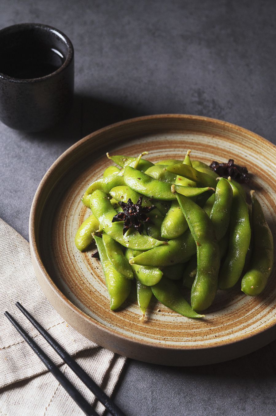 Edamame pods on a tan plate with black sesame seeds, chopsticks, and a cup of tea.