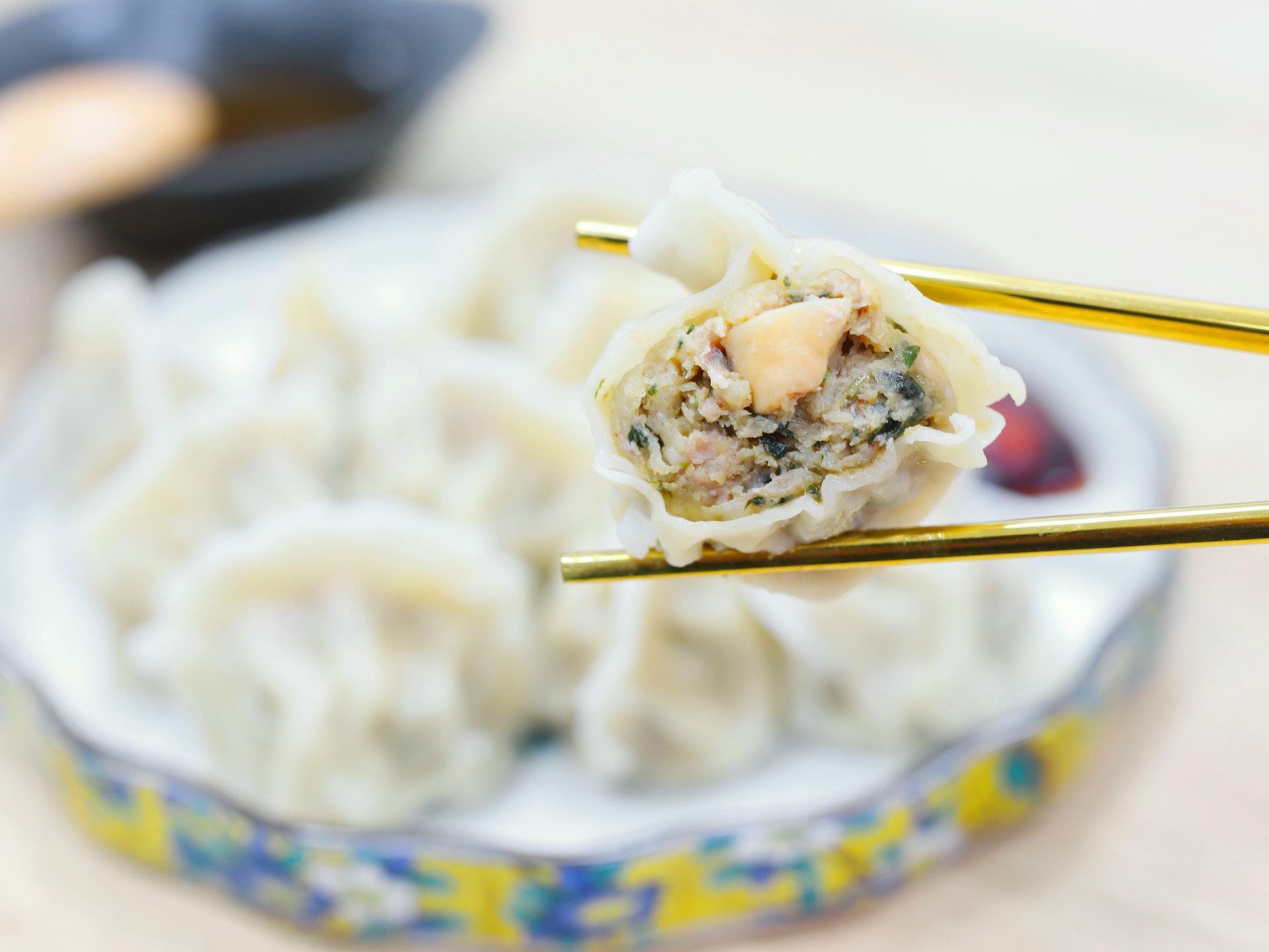 Dumpling held by golden chopsticks, revealing filling; plate of dumplings in background.
