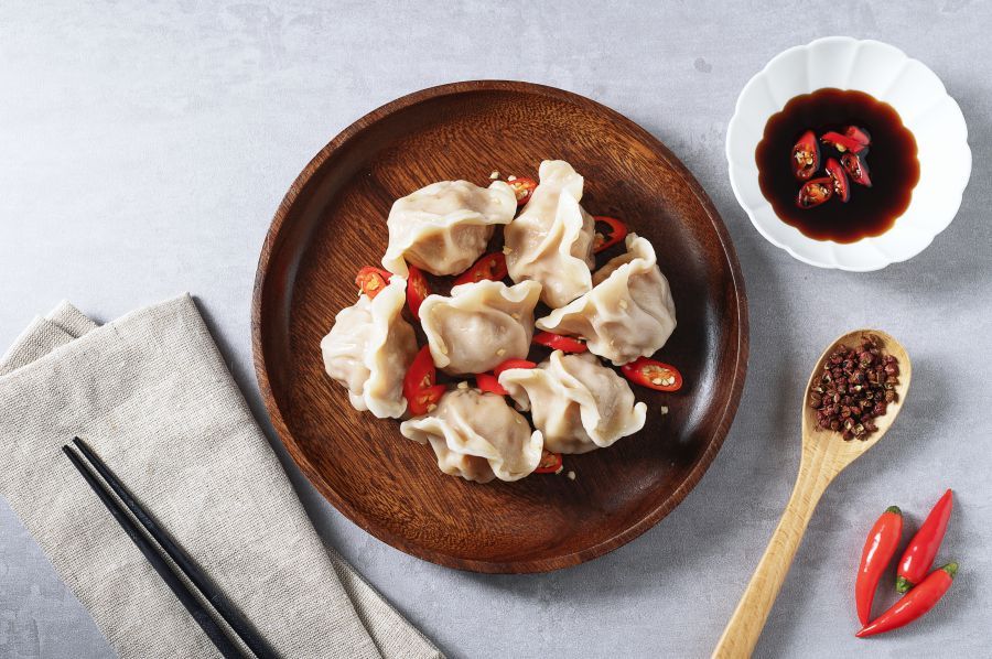 Dumpling held by chopsticks, revealing pork filling, with more dumplings and soy sauce in background.