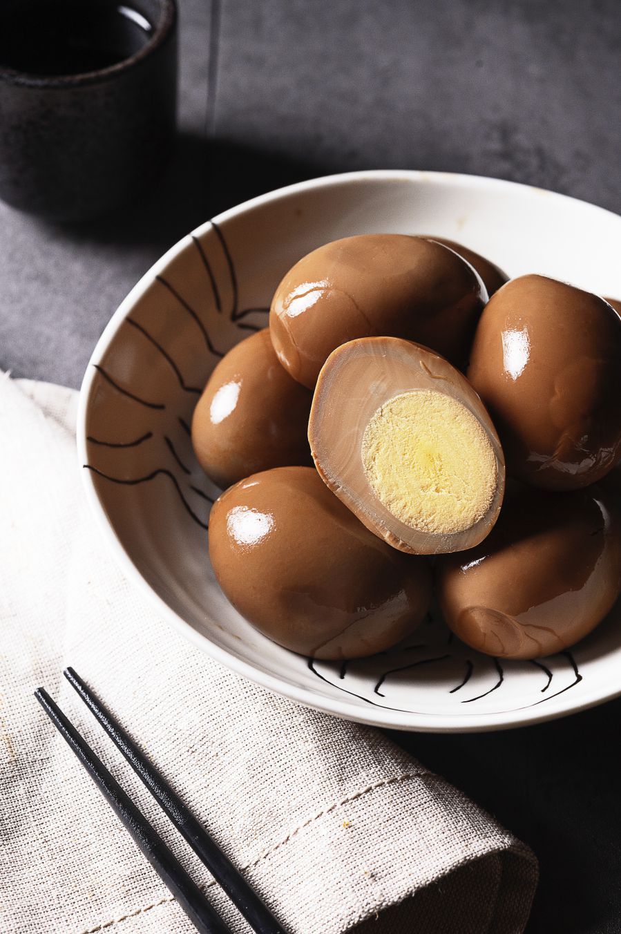 Bowl of brown soy-sauce marinated eggs with a halved egg showing the yellow yolk, served with chopsticks.