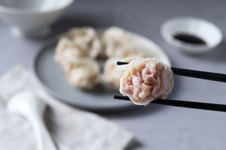 Dim sum held in chopsticks, with a plate of dim sum and dipping sauce in the background.