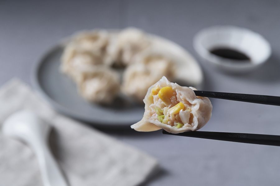 Dumpling held by chopsticks, revealing corn and filling. Plate with more dumplings and soy sauce.