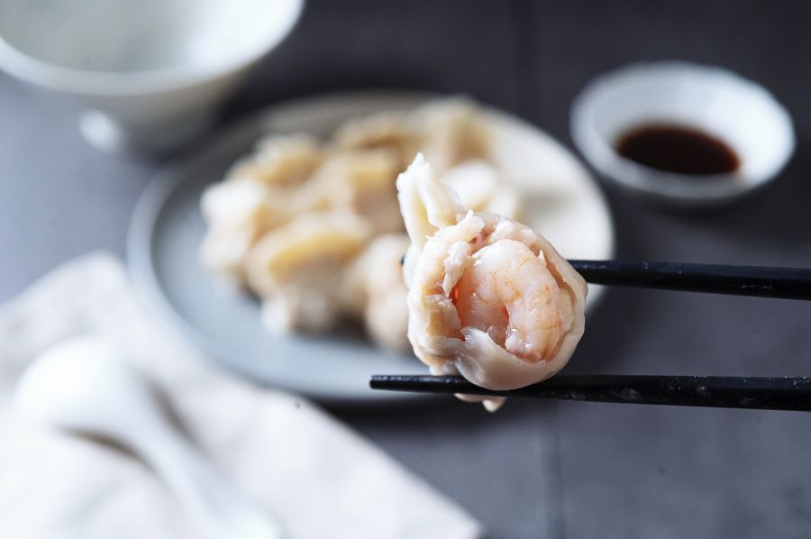 Shrimp dumpling held by black chopsticks, plate of dumplings, small dipping sauce, grey background.
