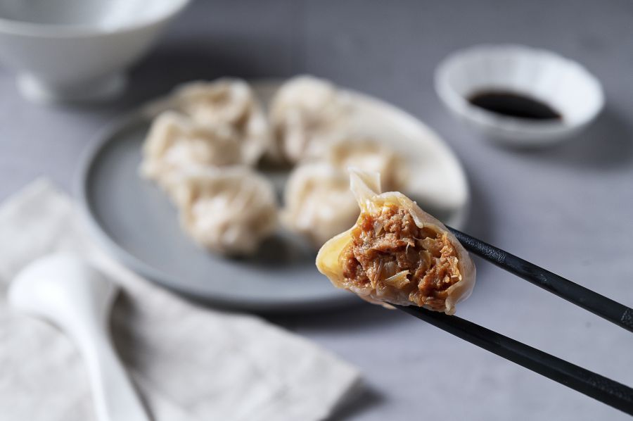 Dumpling held by chopsticks, revealing pork filling, with more dumplings and soy sauce in background.