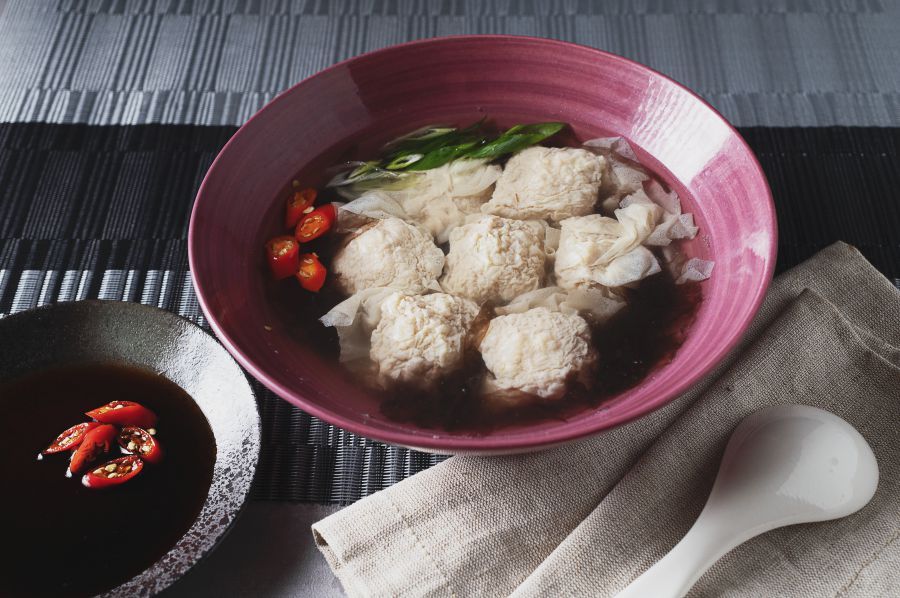 Soup with meatballs, vegetables, and sauce in a bowl. A spoon and sauce dish are beside it.
