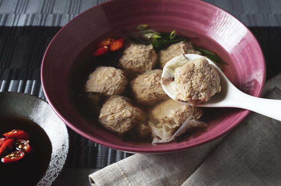 Meatballs in broth, served in a pink bowl with a spoon. Red chili peppers and green onions add color.