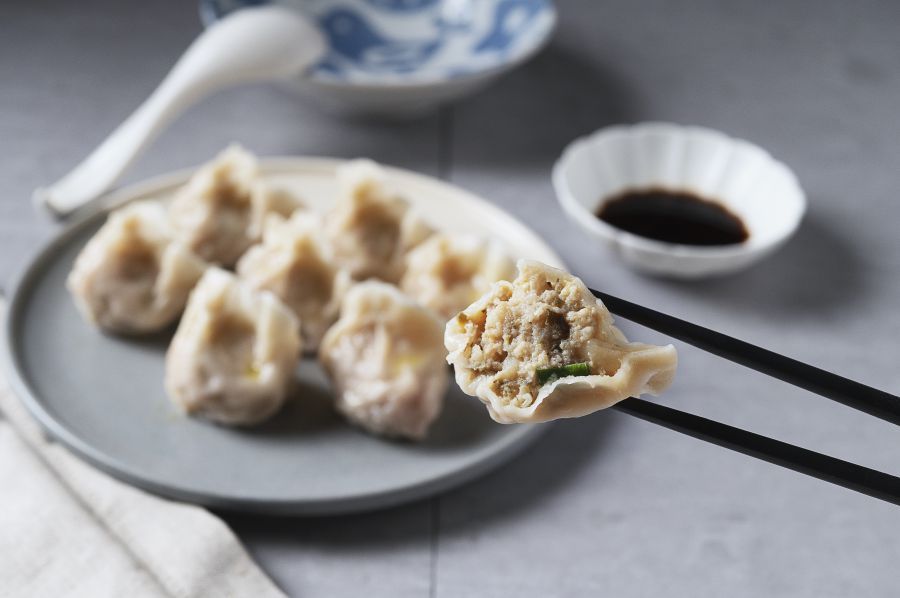 Dumplings on plate, held by chopsticks, with soy sauce and spoon on a table.