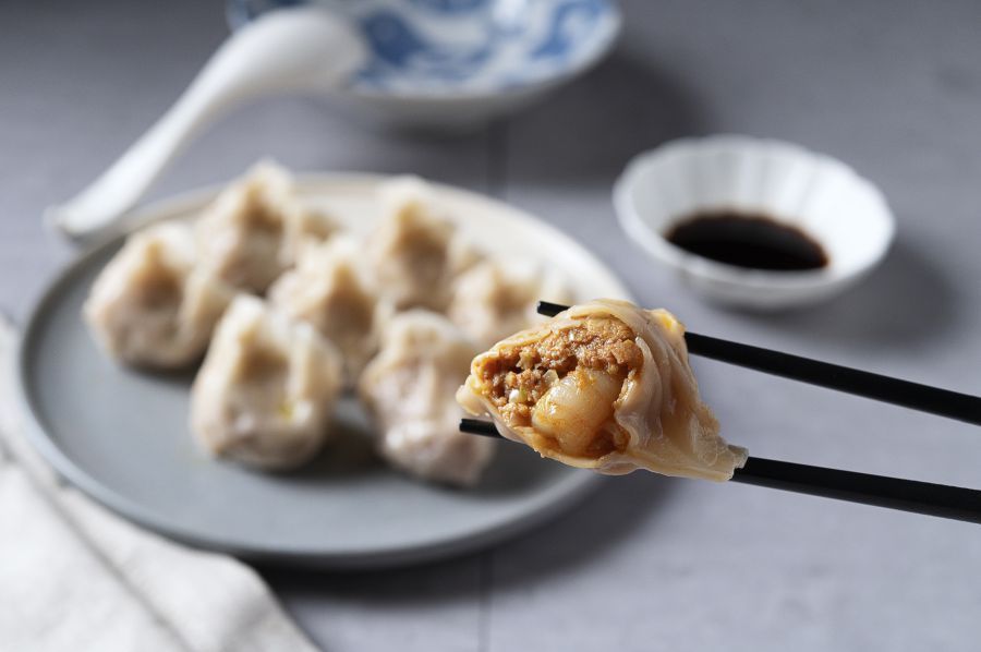 Dumplings on a plate, one being held by chopsticks with dipping sauce, ceramic bowl, and spoon in the background.