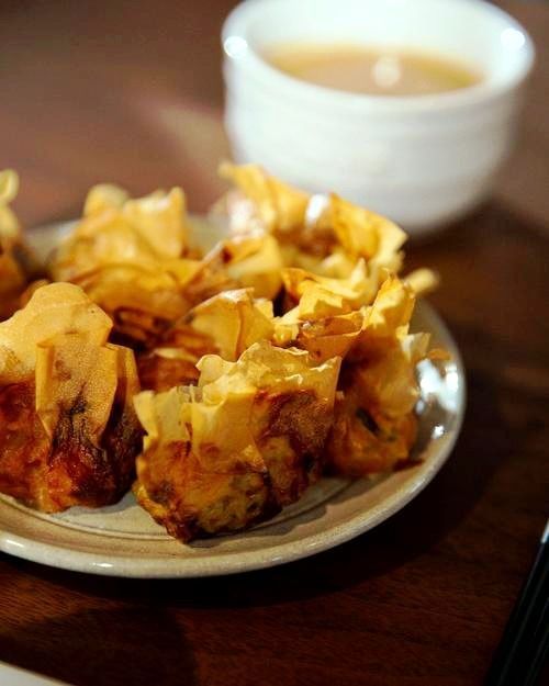 Plate of golden fried wontons, near a cup of liquid, on a wooden surface.