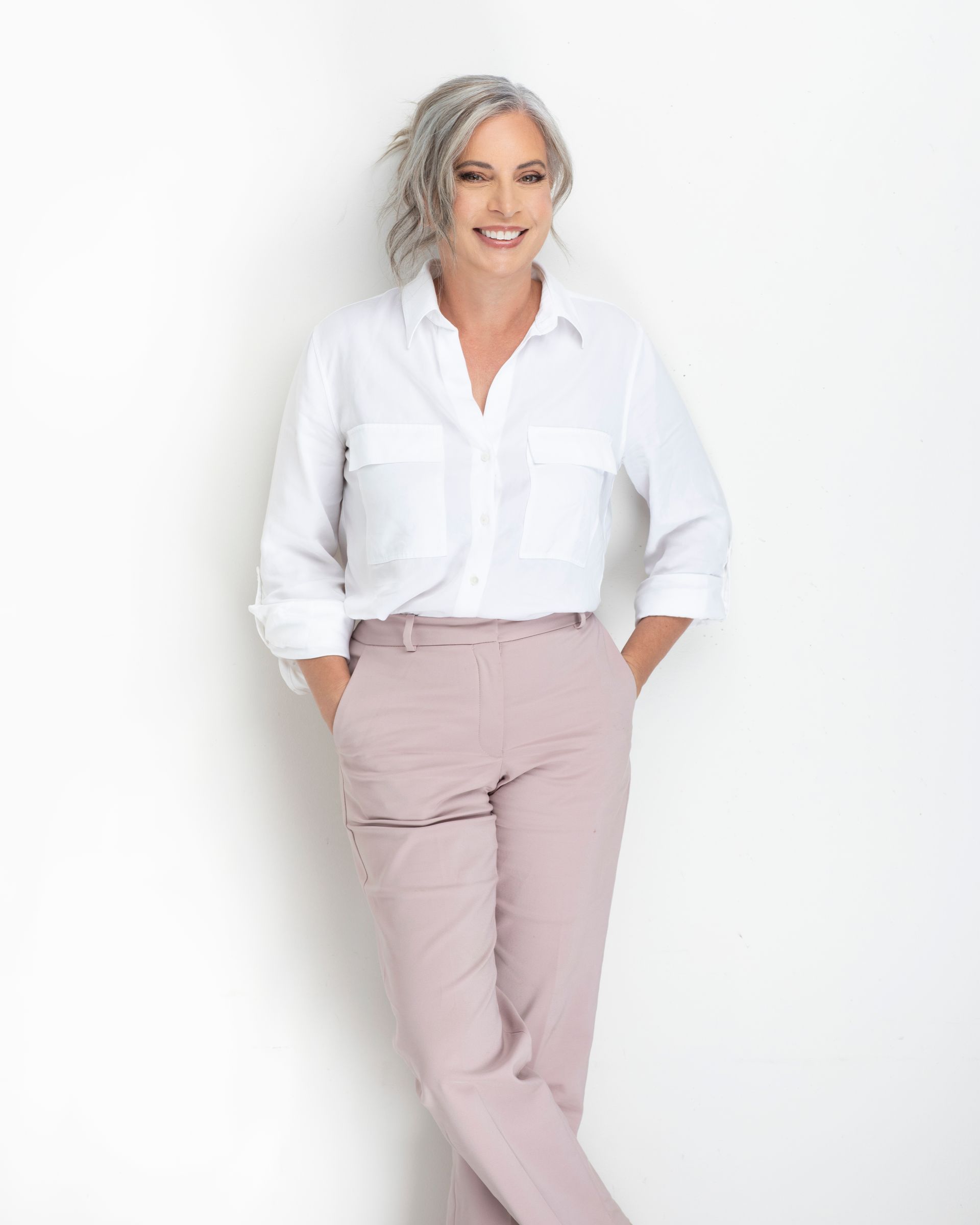 A photo of Michelle Andreacchi standing in front of a white background with a white blouse and light pink dress pants.