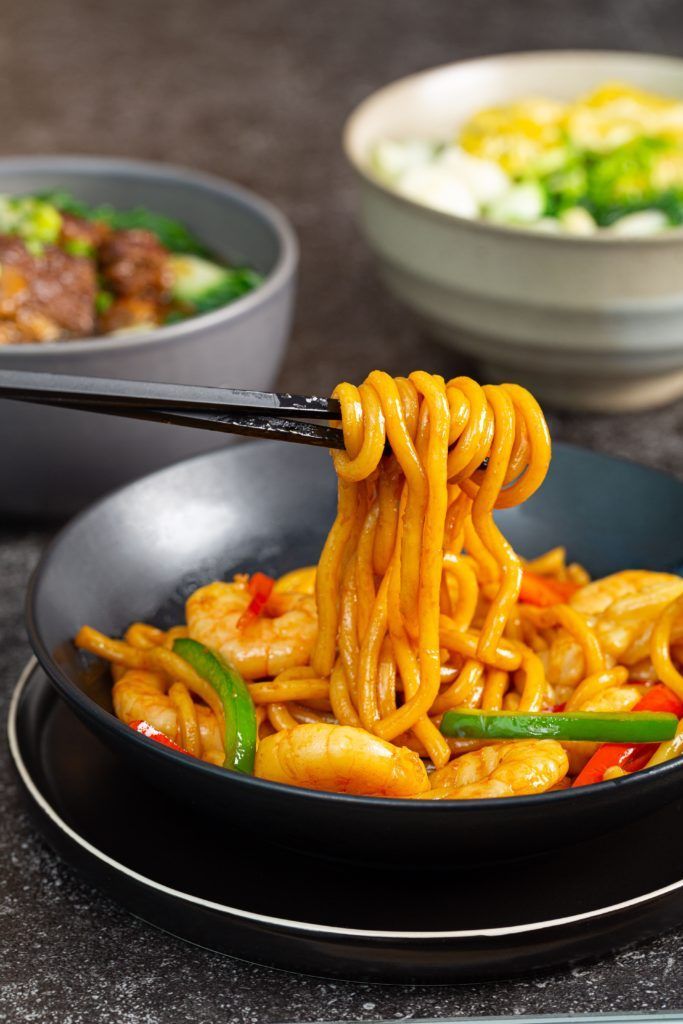 A bowl of noodles with shrimp and vegetables is being eaten with chopsticks.