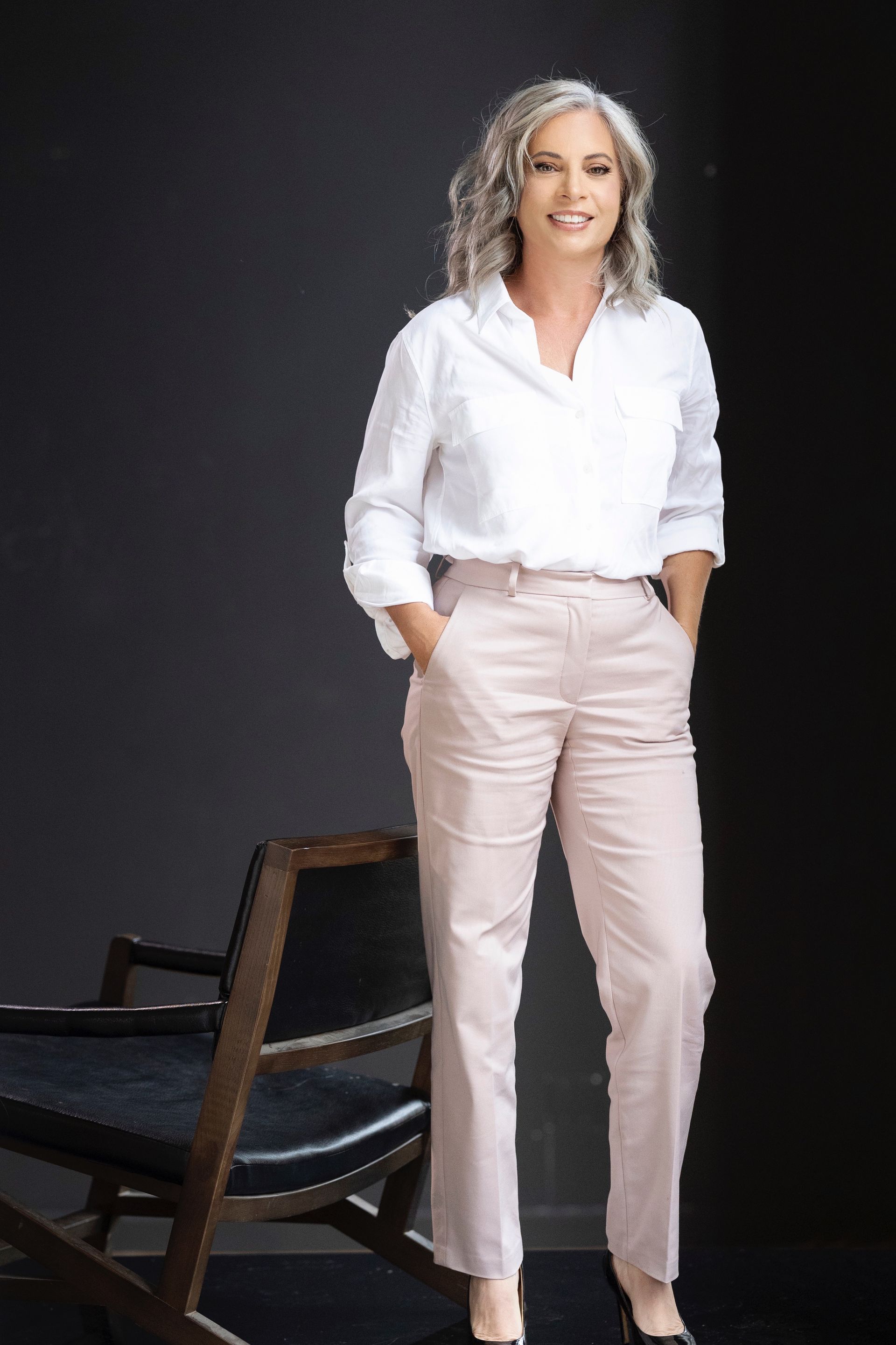 A woman in a white shirt and pink pants is standing next to a chair.