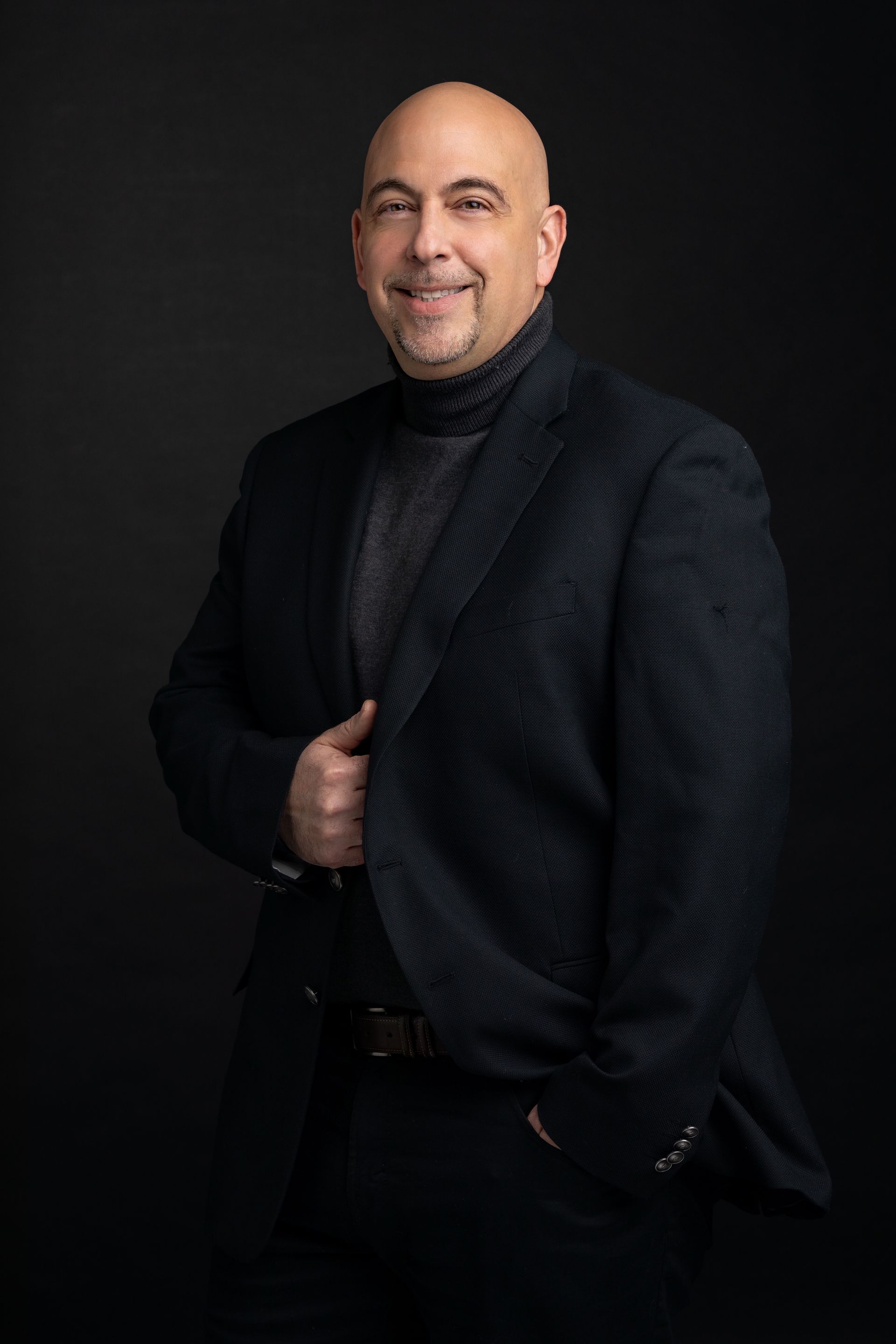 A man in a suit and turtleneck is standing in front of a black background.