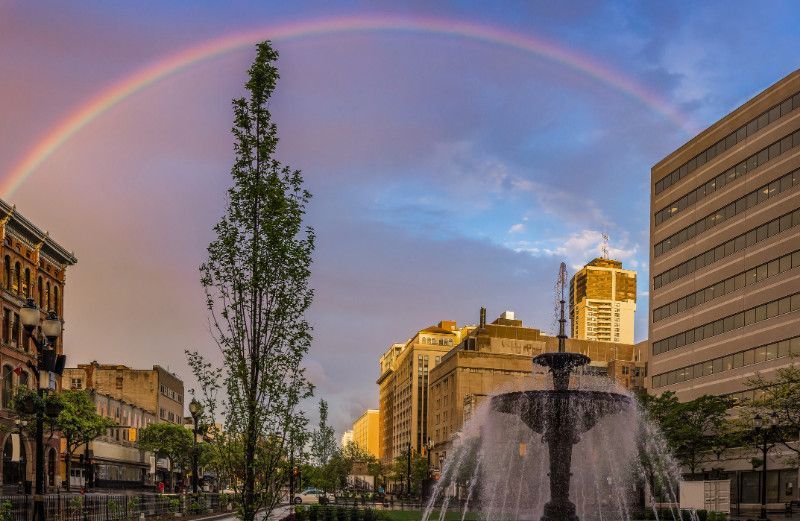 There is a rainbow over a fountain in the middle of a city.