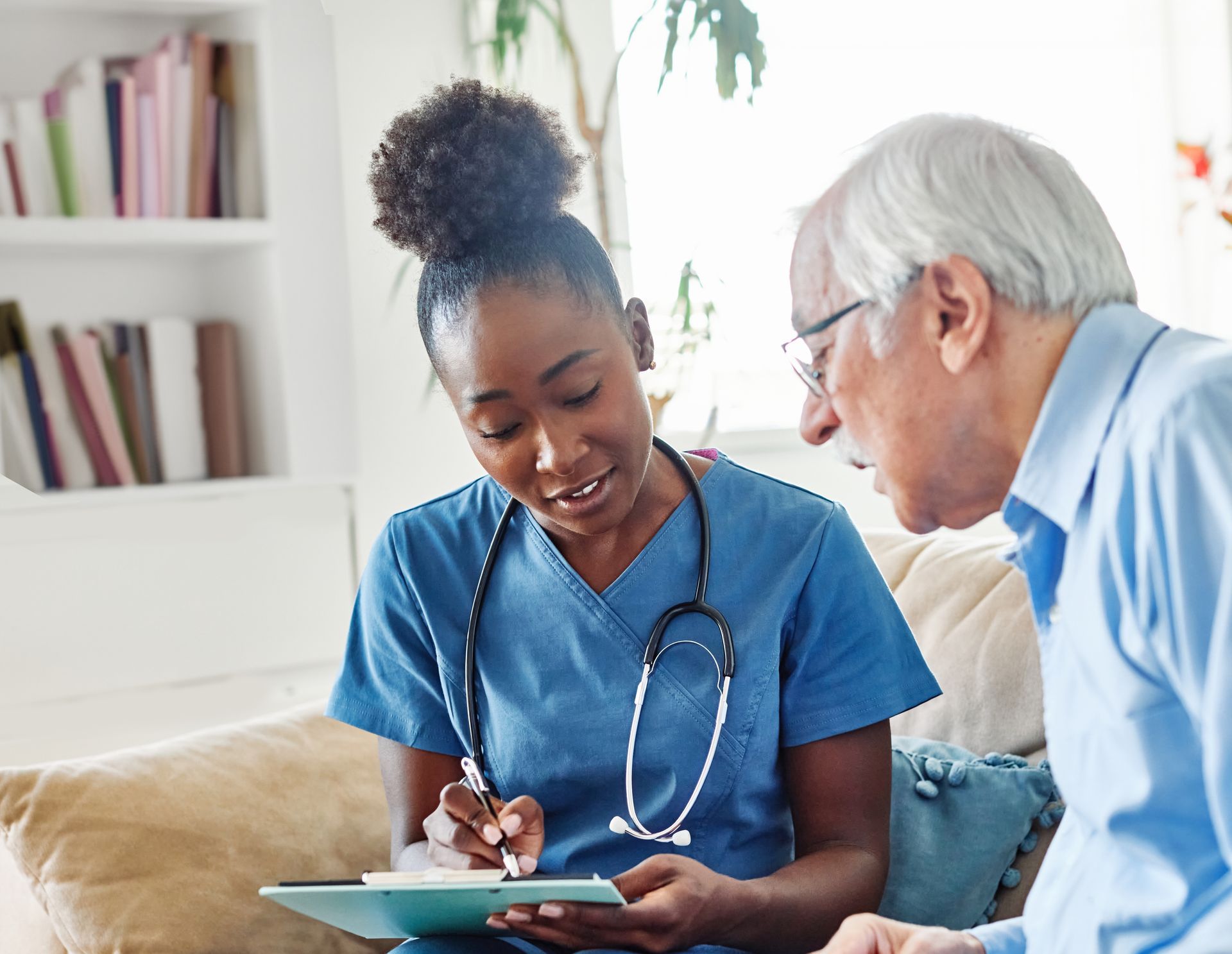 Caregiver points to a notebook with elderly woman, both smiling, sitting on a sofa.