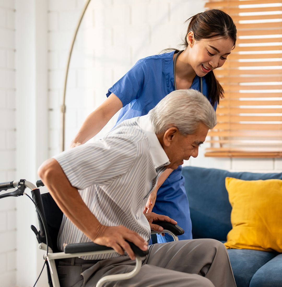 Caregiver assists an older adult from wheelchair. They are indoors with a blue couch.