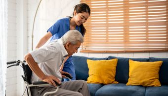 Caregiver assists an older adult from wheelchair to couch. Indoors, blue couch, yellow pillows.