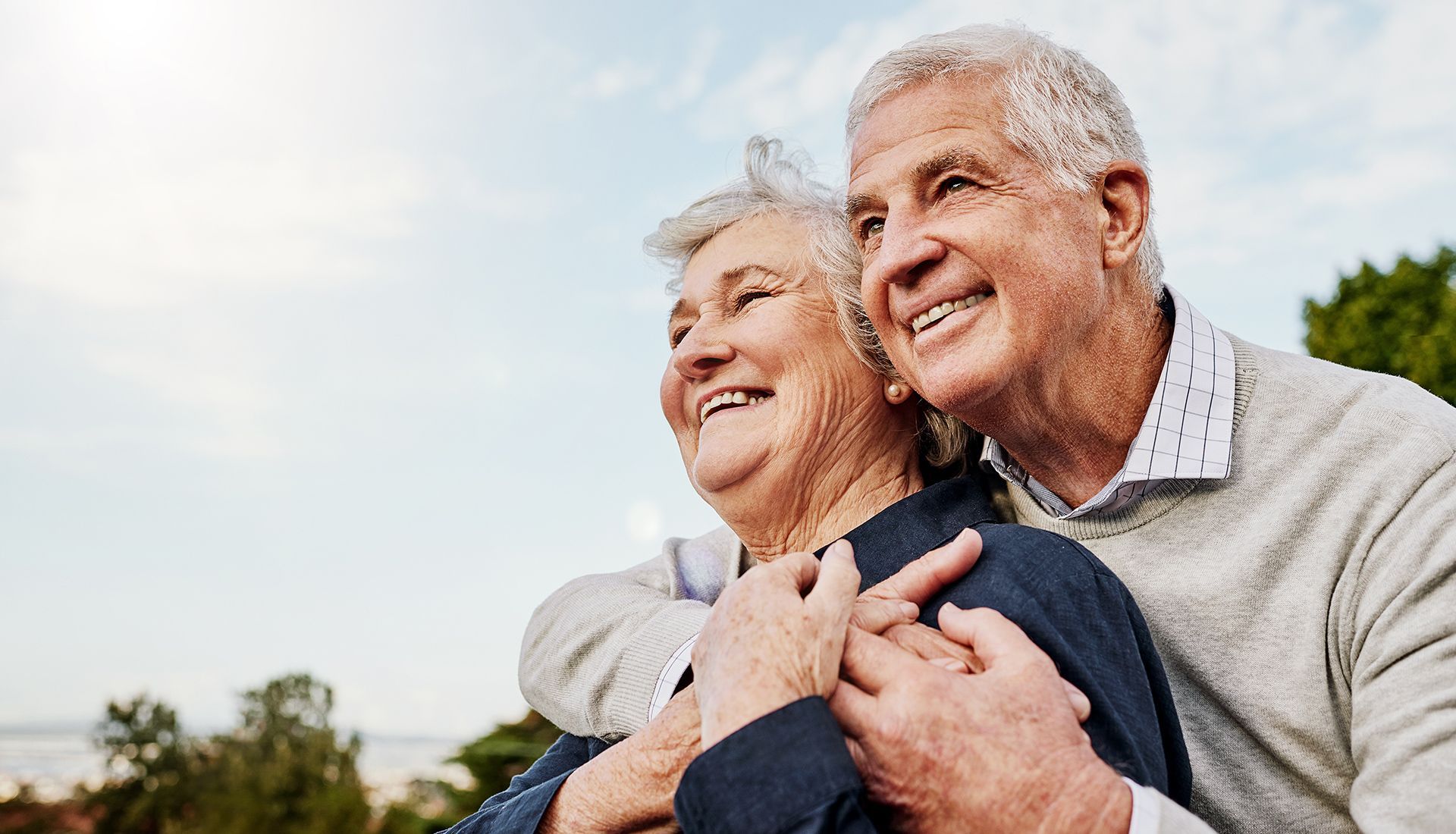Elderly couple embraces and smiles outdoors, under a blue sky.