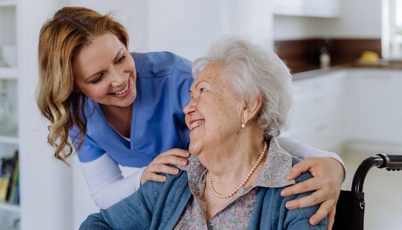 Woman in blue scrubs smiles while assisting elderly person seated in a wheelchair indoors.