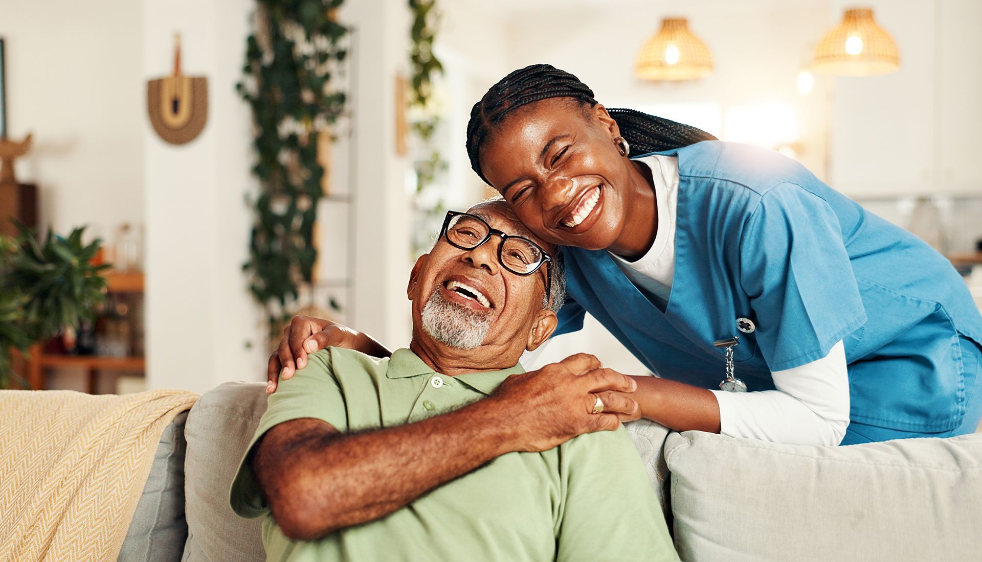 Woman in scrubs embraces smiling elderly person on a couch in a home setting.