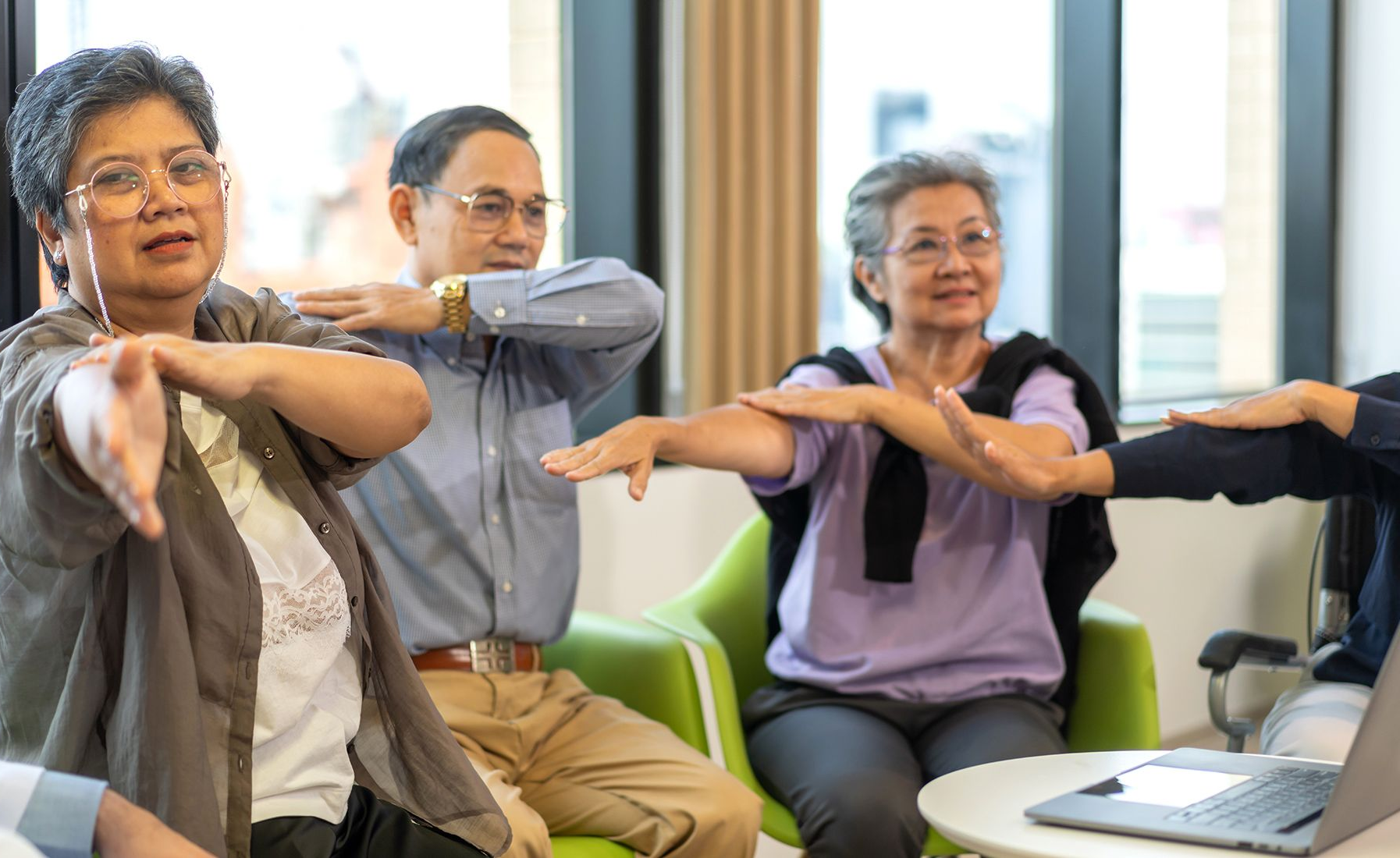 Three older adults stretching arms, possibly exercising in a well-lit room.