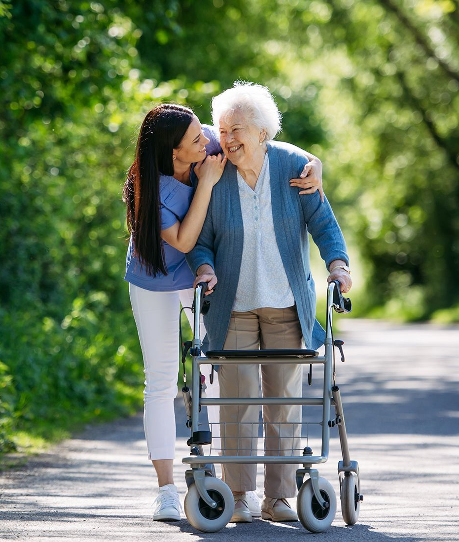 Woman assisting elderly person using a walker on a path, both smiling.