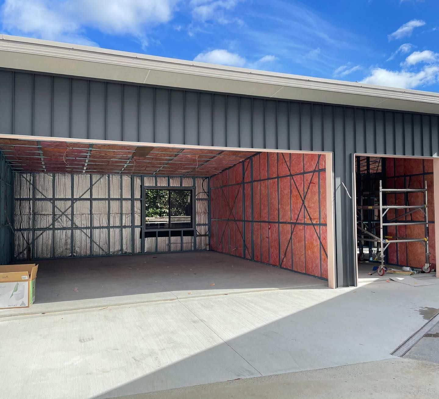 Exterior view of a garage under construction, with open bays and exposed insulation. Gray siding and concrete floor.