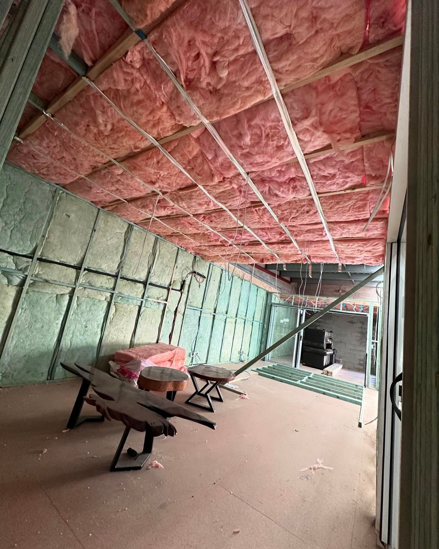Interior of a room under construction with pink and green insulation on walls and ceiling; two wooden benches sit inside.