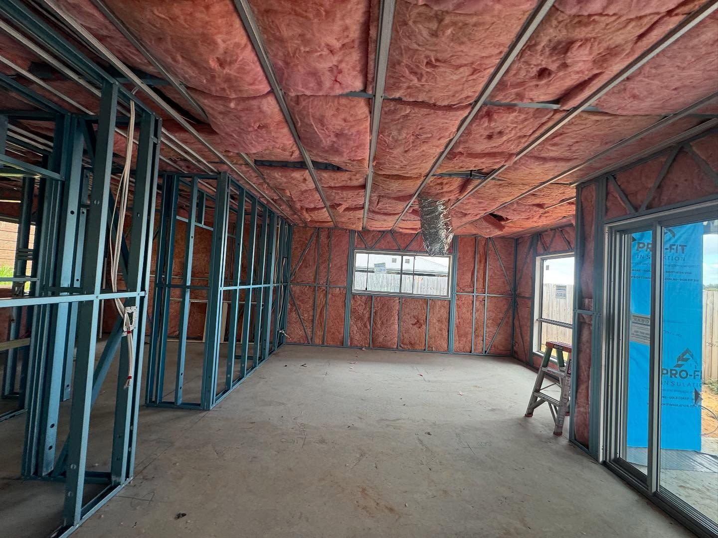 Interior of a room under construction with metal framing, pink insulation, windows, and a concrete floor.