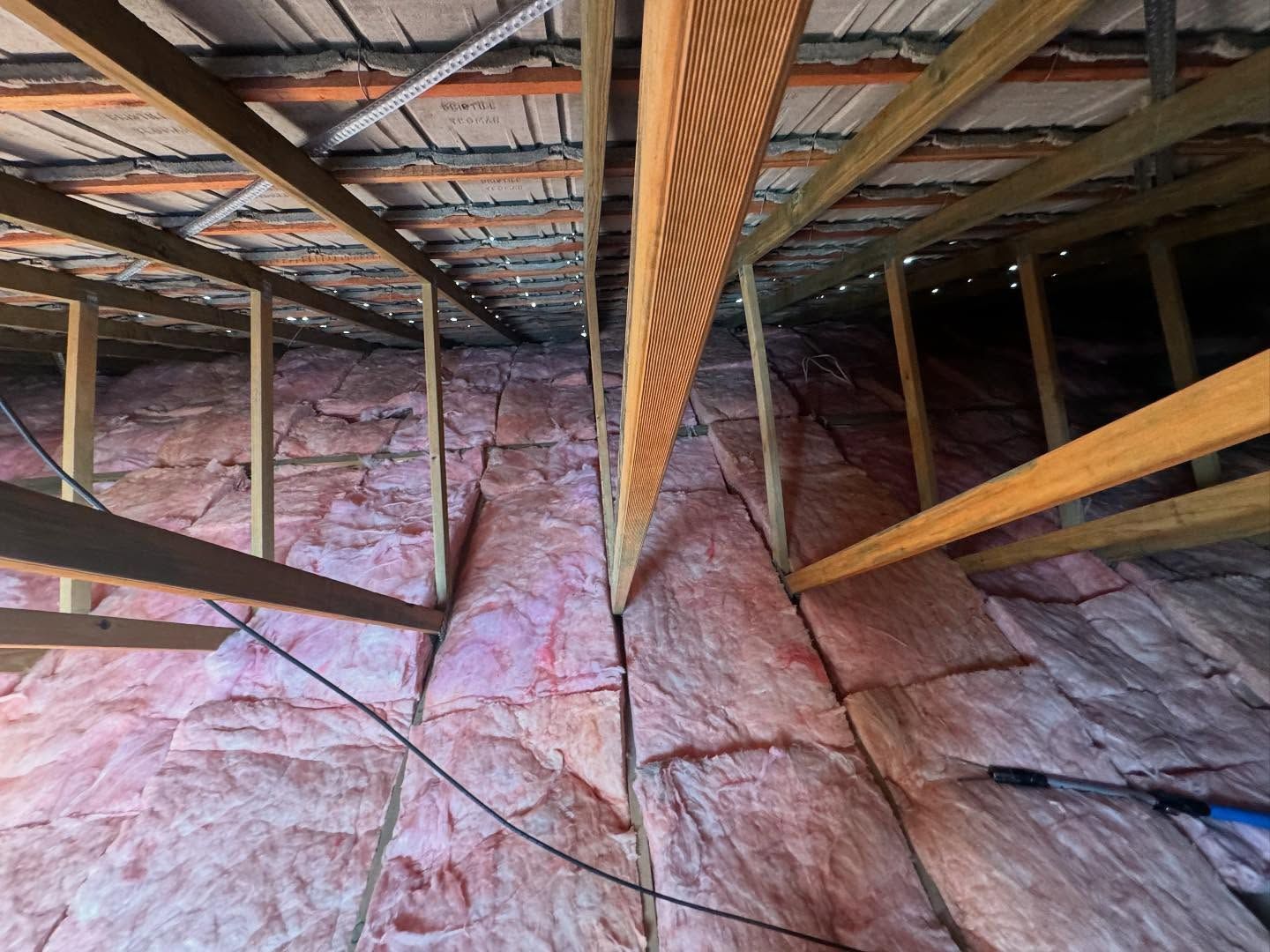 Pink insulation lines the interior of an attic, with wooden beams and rafters overhead.