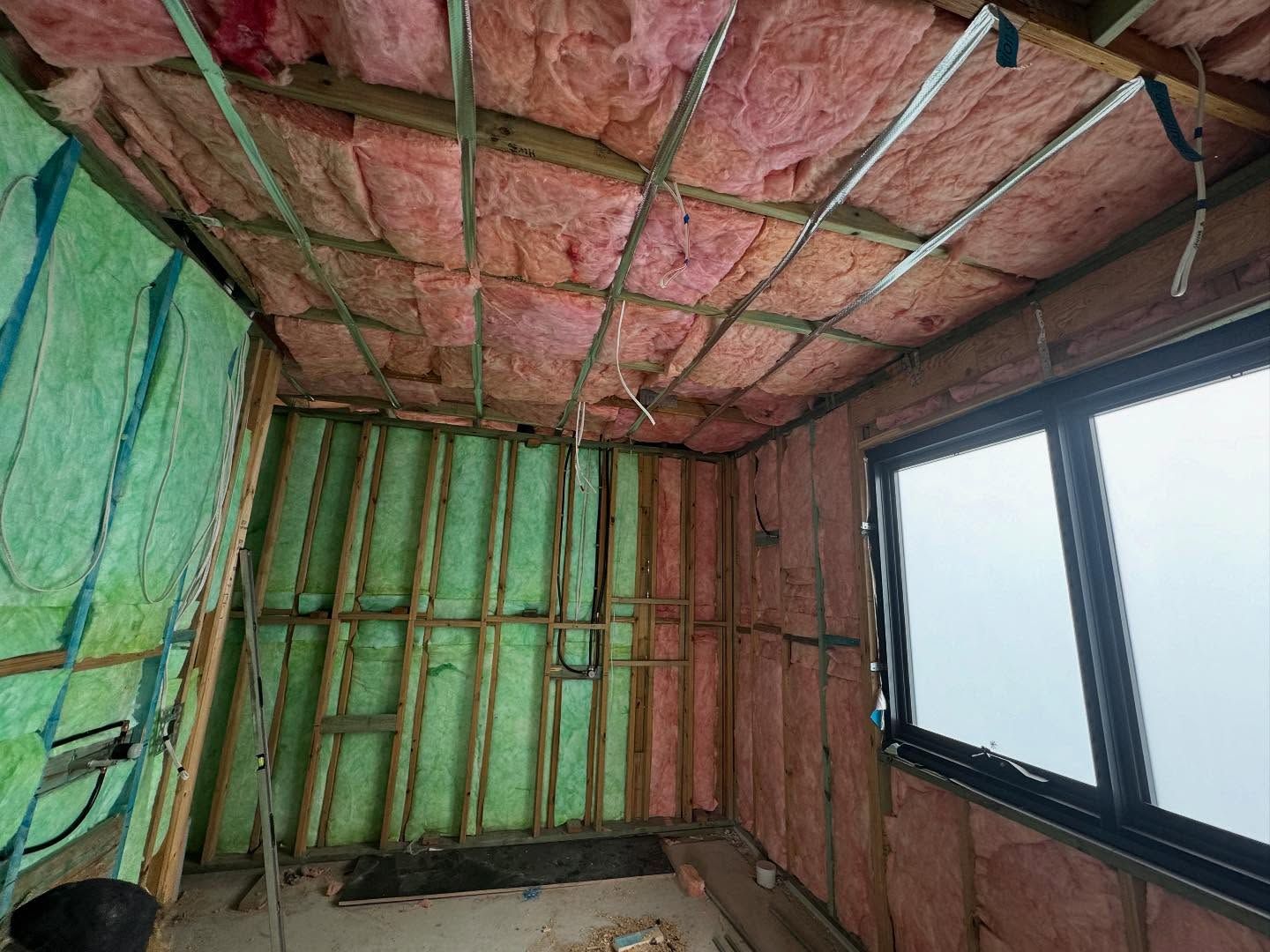 Interior of a room under construction, with pink insulation in the ceiling and walls. A window is visible.