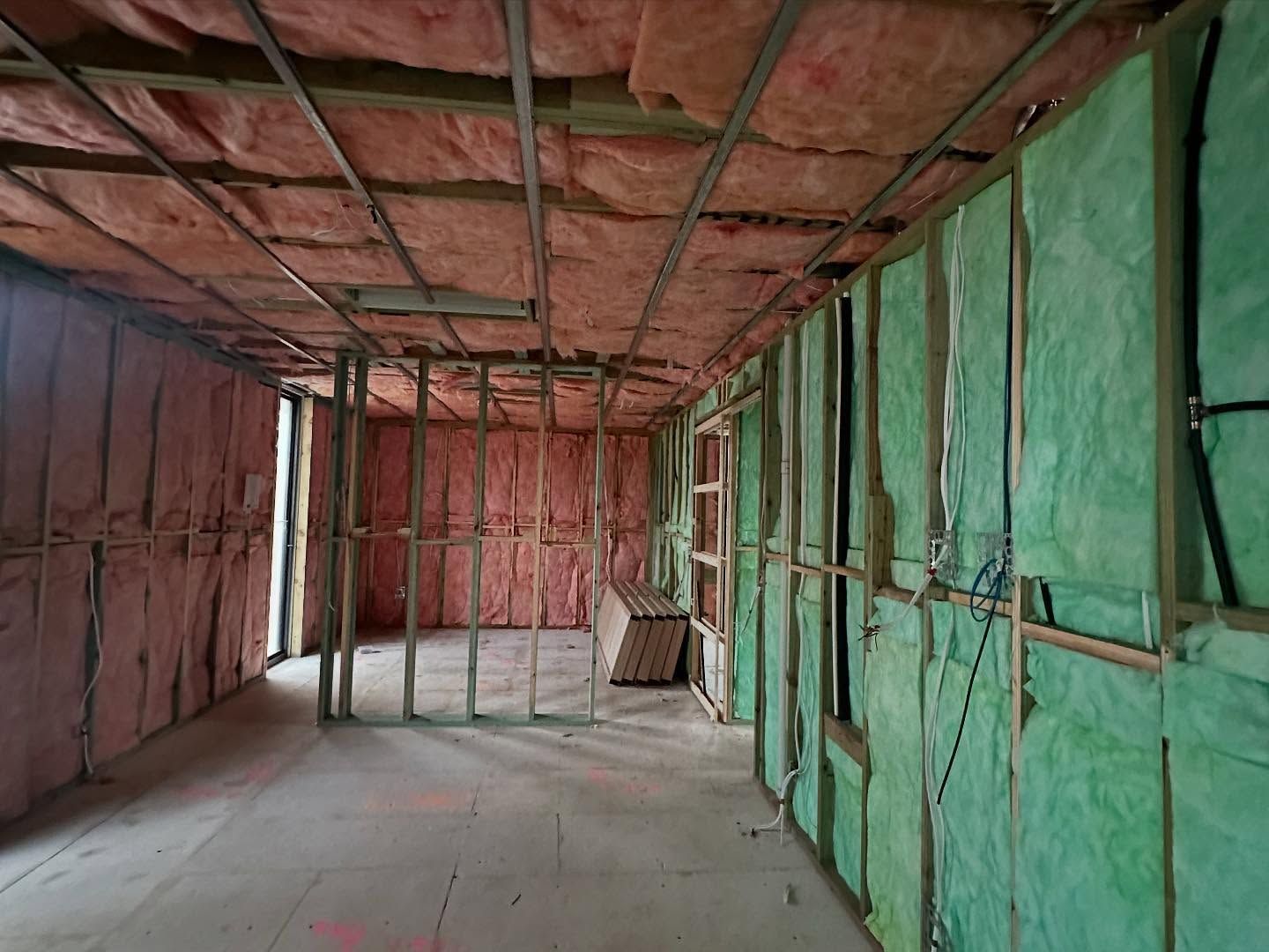 Interior view of a room under construction, showing insulation in pink and green, wood framing, and metal grid ceiling.
