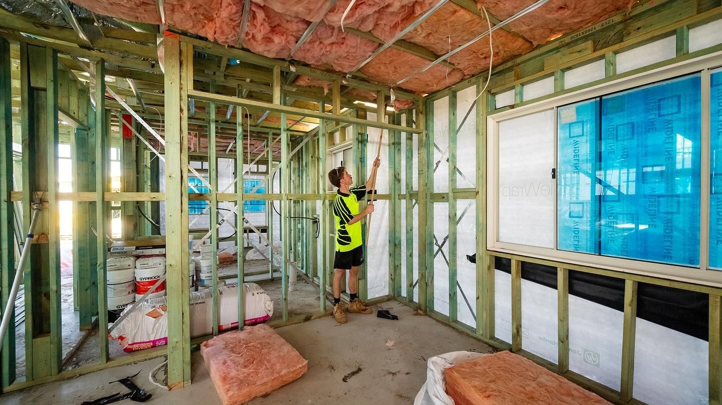 Interior framing of a building under construction, a worker in a safety vest reaching towards a ceiling wire.