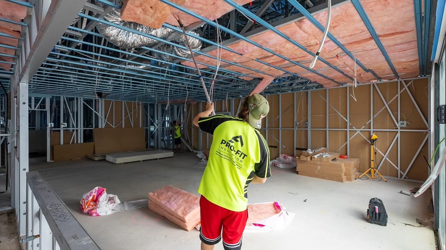 Construction worker installing insulation in a room under construction, wearing a yellow shirt and red shorts.