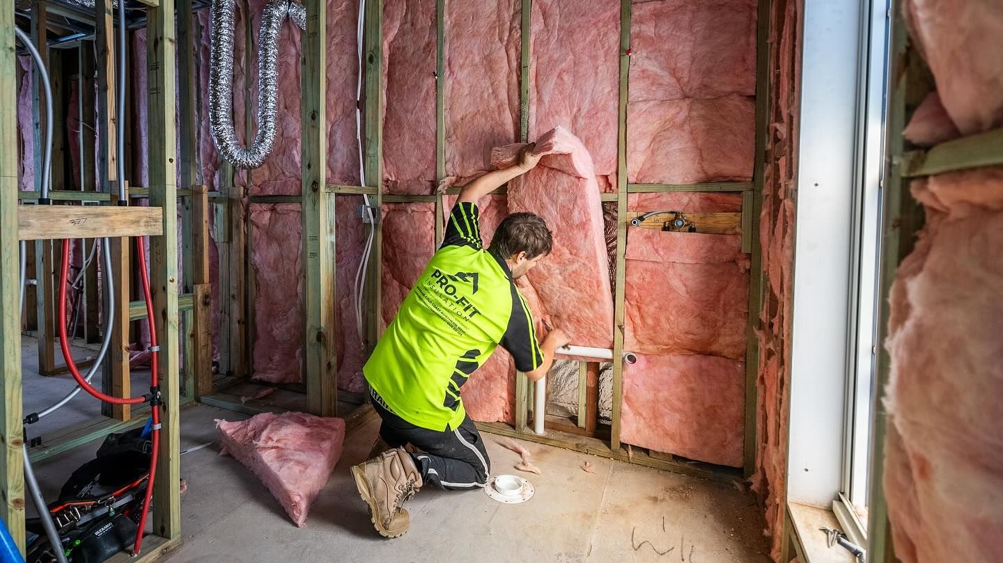 Construction worker installing pink insulation in a partially built room, kneeling.