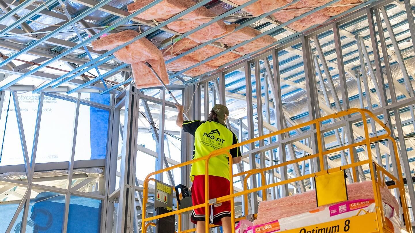 Construction worker installing pink insulation in a building's ceiling, standing on a lift.