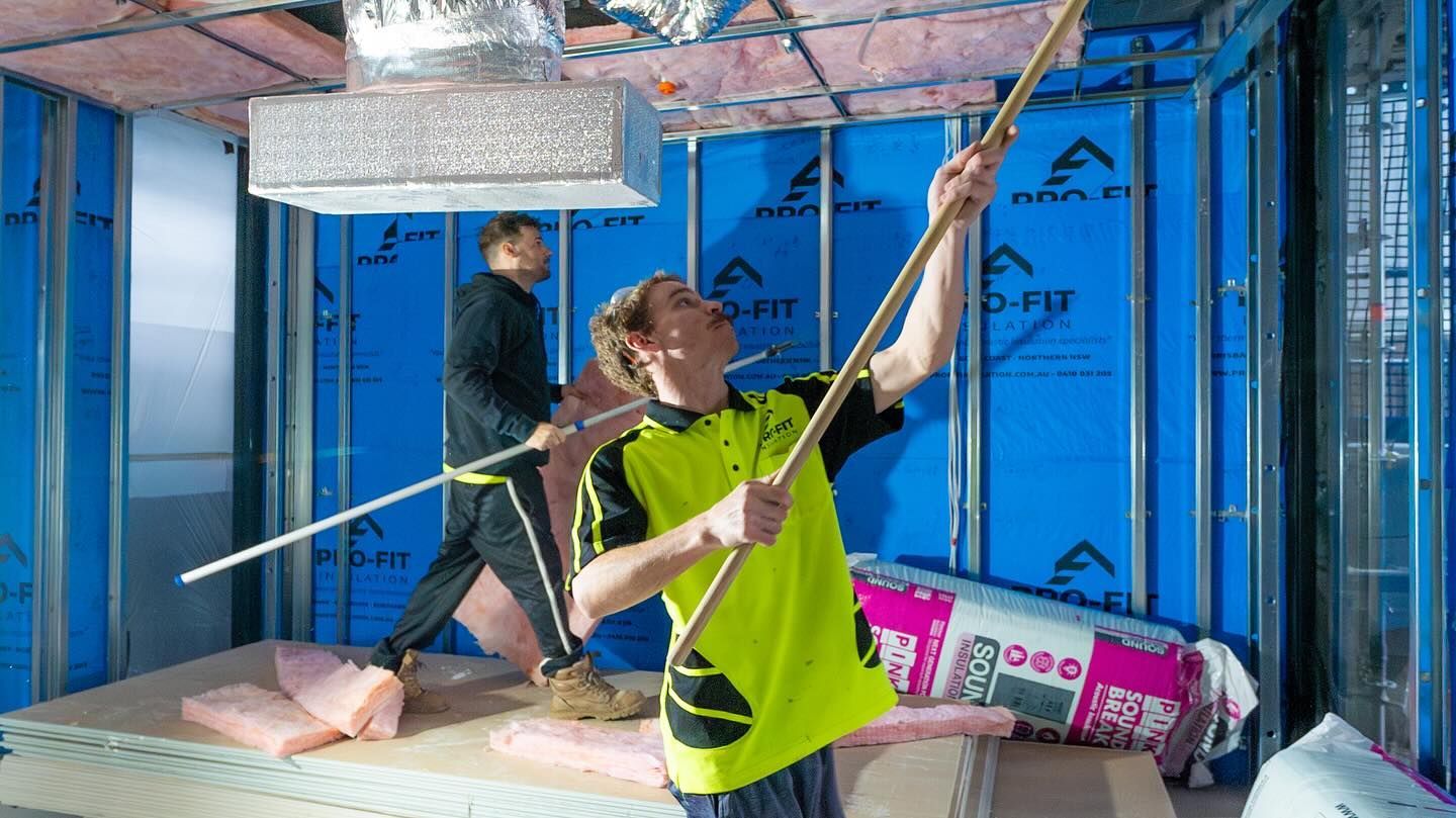 Two workers installing insulation in a room with blue walls, metal studs, and ductwork. One uses a long tool.