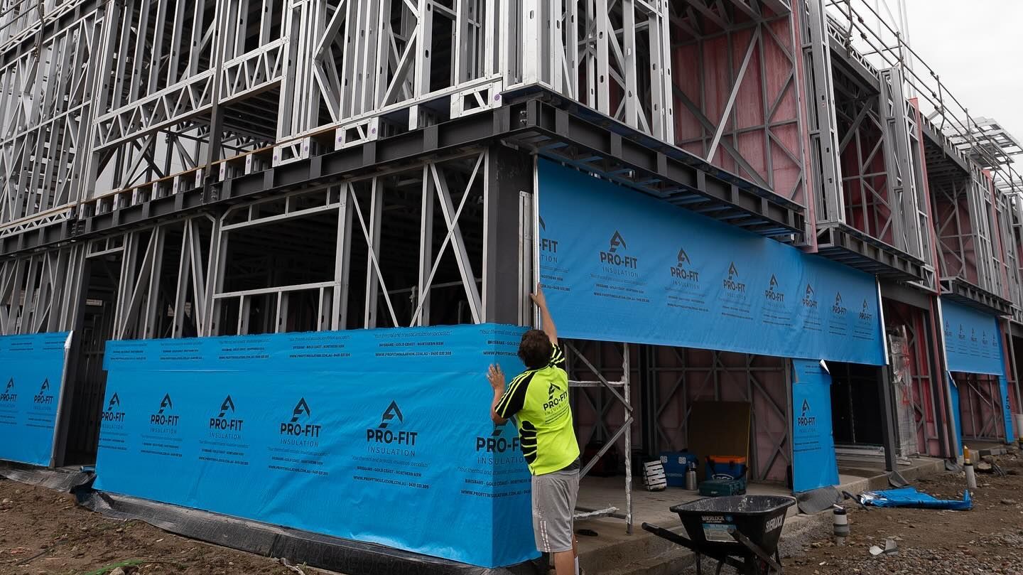 Construction worker installing blue weather barrier on a steel-framed building.