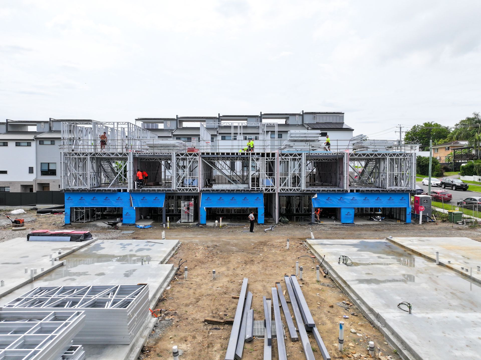 Construction site with multi-unit buildings under construction. Steel framing, concrete foundations, and workers visible.
