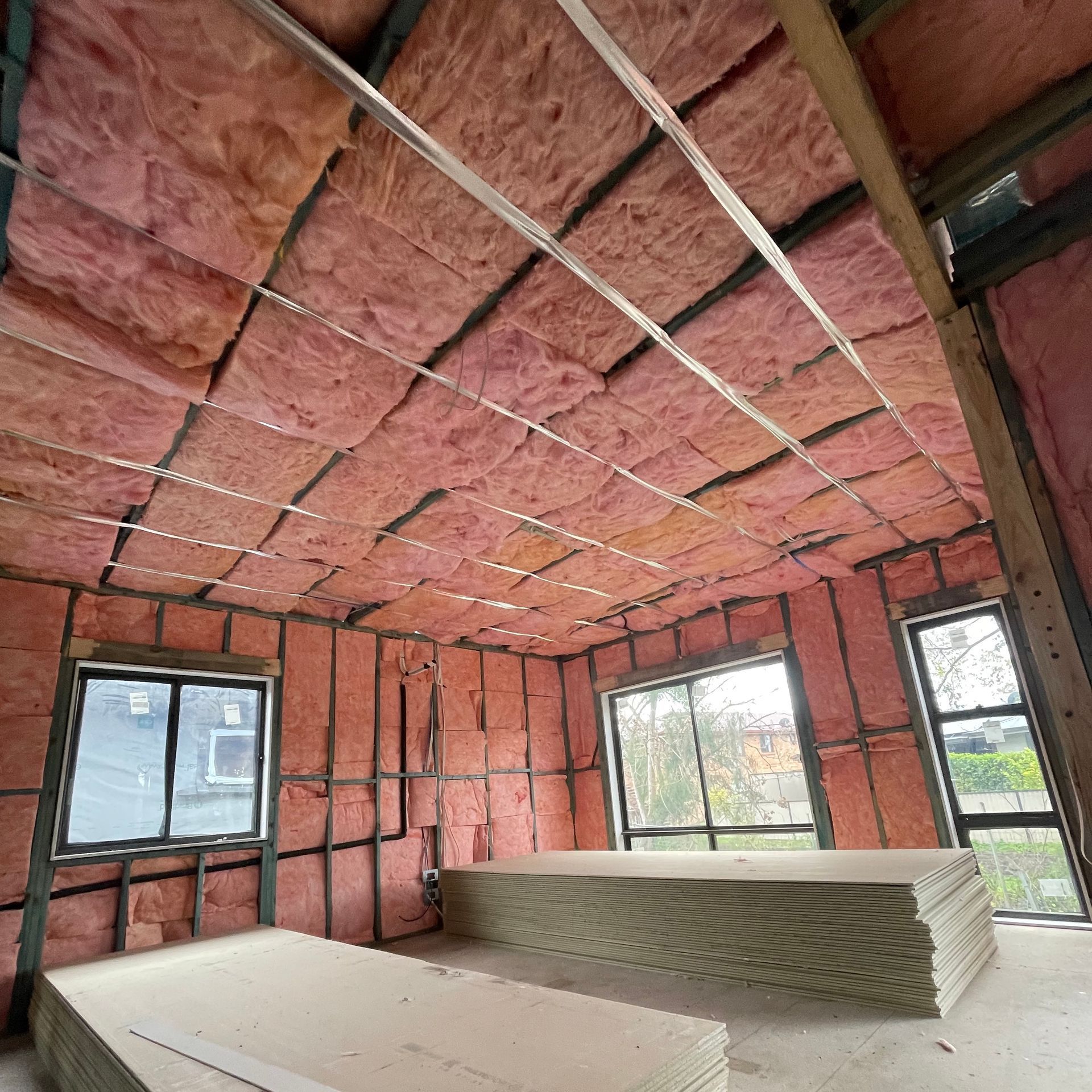 Interior room under construction, with pink insulation on the walls and ceiling, and stacks of drywall on the floor.