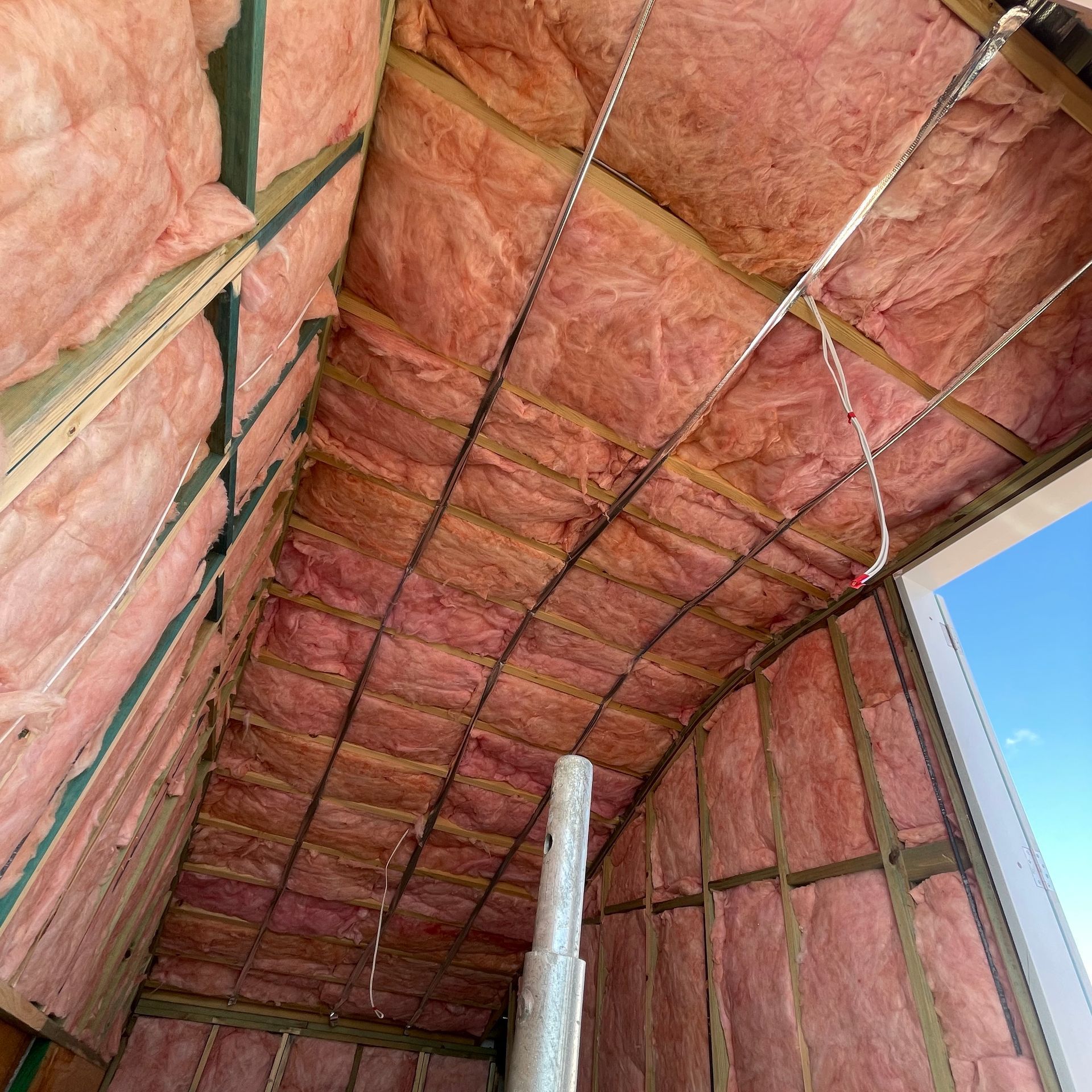Pink insulation installed in an attic with wooden beams and a white PVC pipe.