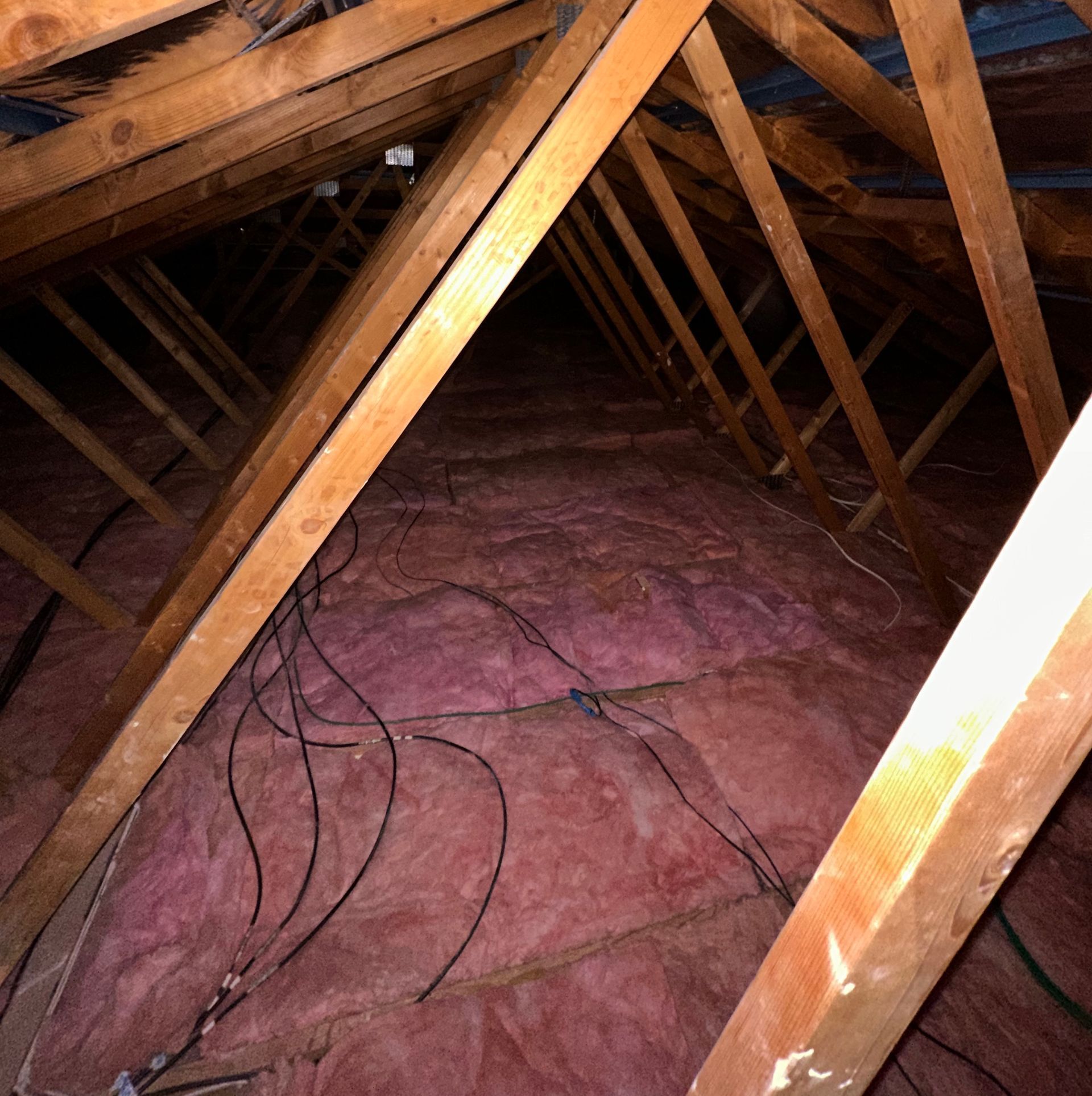 View from inside an attic. Wooden rafters support a pink insulation layer, with black wires running through.