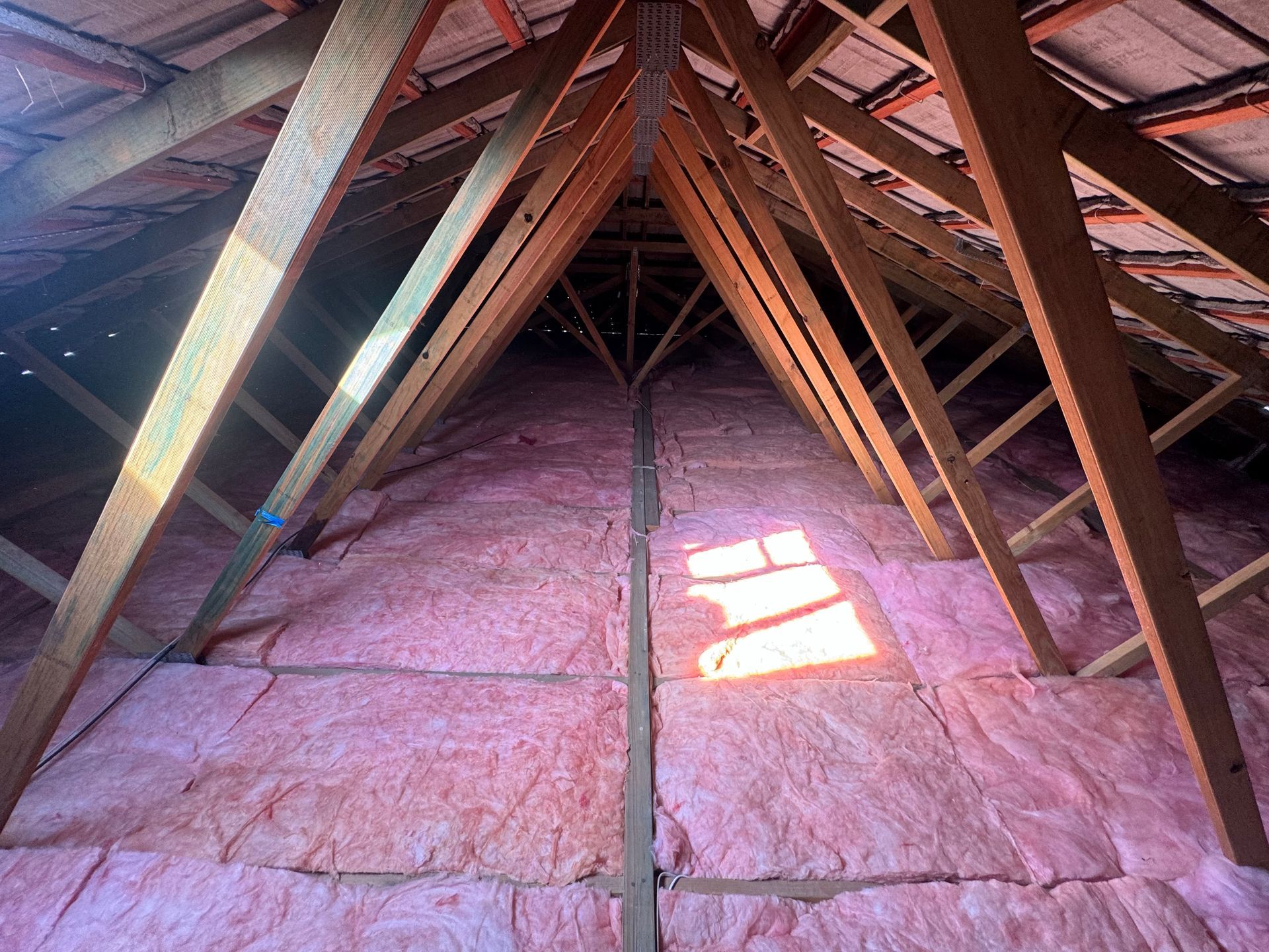Interior view of an attic with pink insulation between wooden rafters.