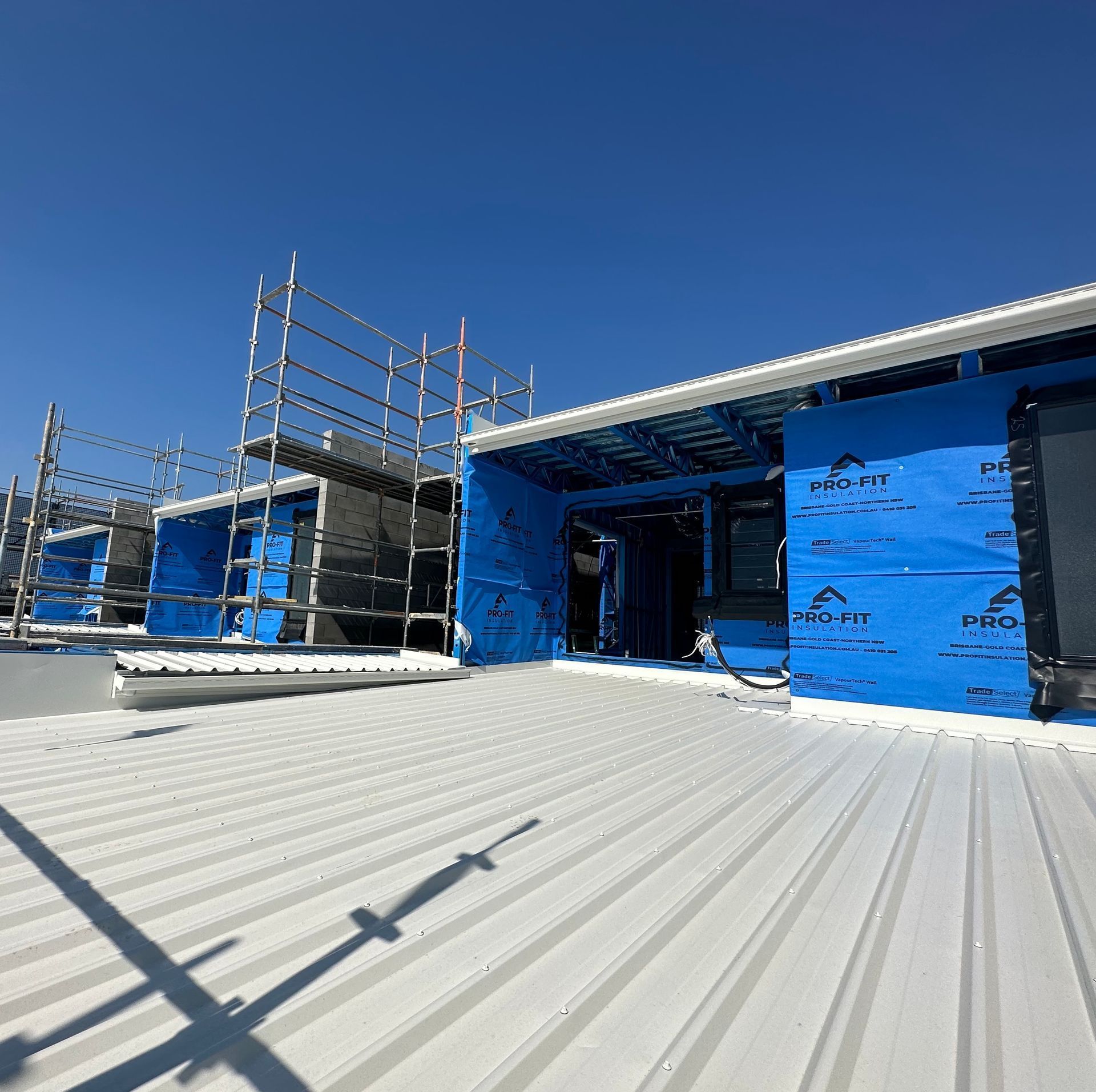 A roof construction site on a sunny day. Scaffolding, blue wrap, and metal roofing are visible.
