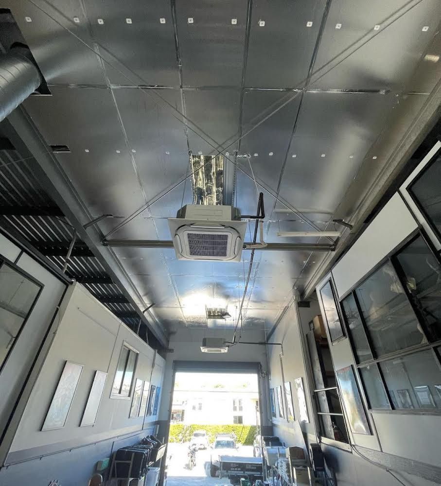 A narrow industrial space with a shiny ceiling and a device hanging overhead, garage door open to the outdoors.