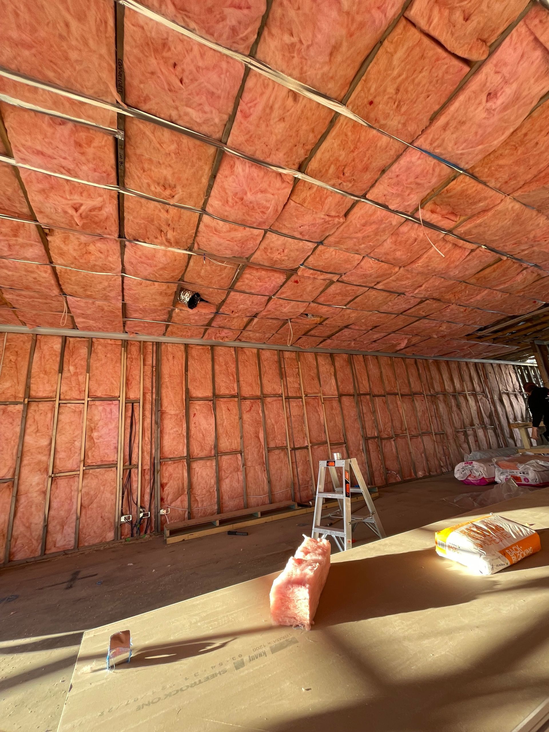 Interior view of a room under construction, insulated with pink fiberglass batting. A ladder and materials are present.