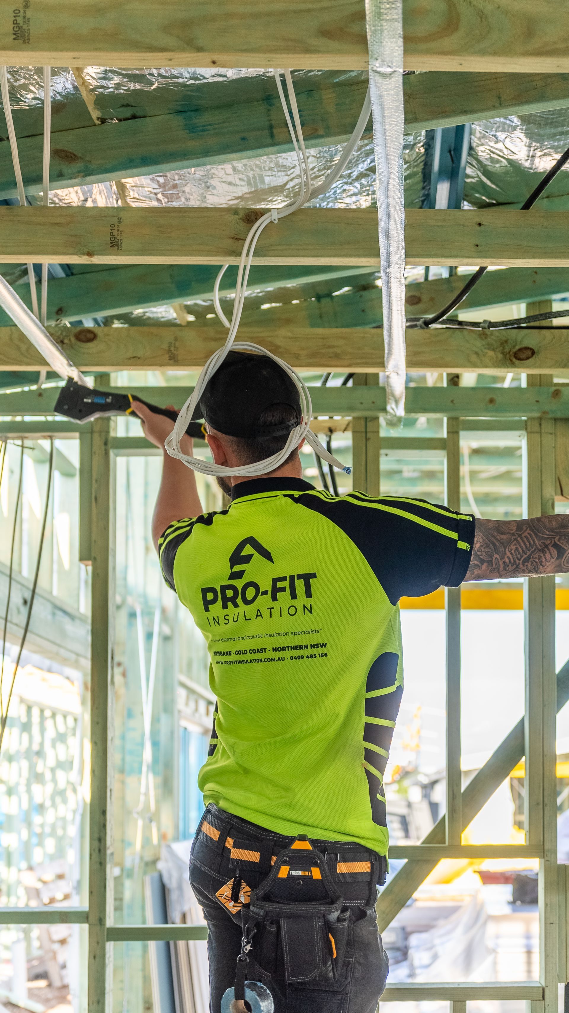 Construction worker in a lime green shirt with tool belt, drilling into wooden ceiling beams.