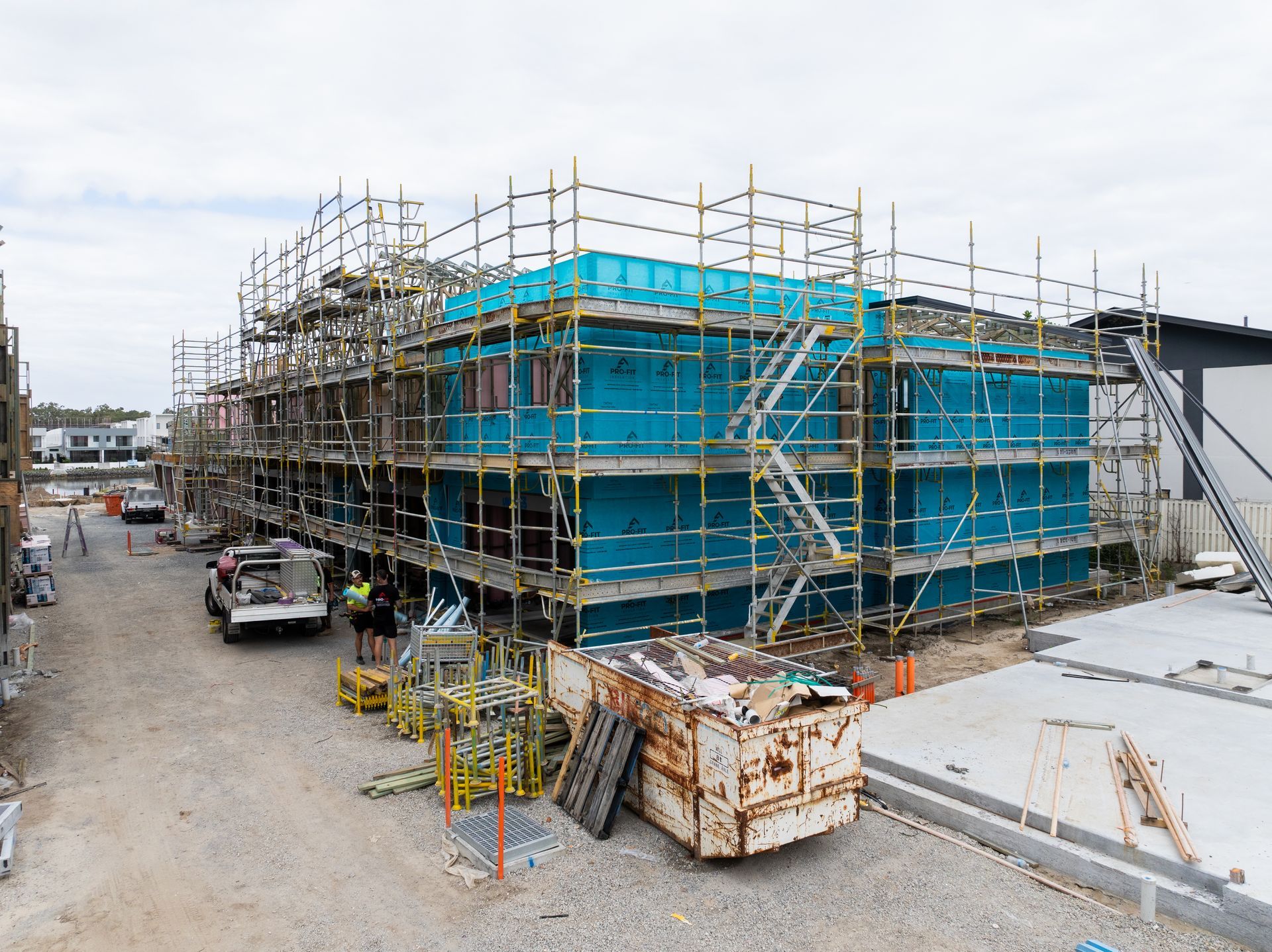 Building under construction with scaffolding, teal exterior, concrete path, and waste bin.